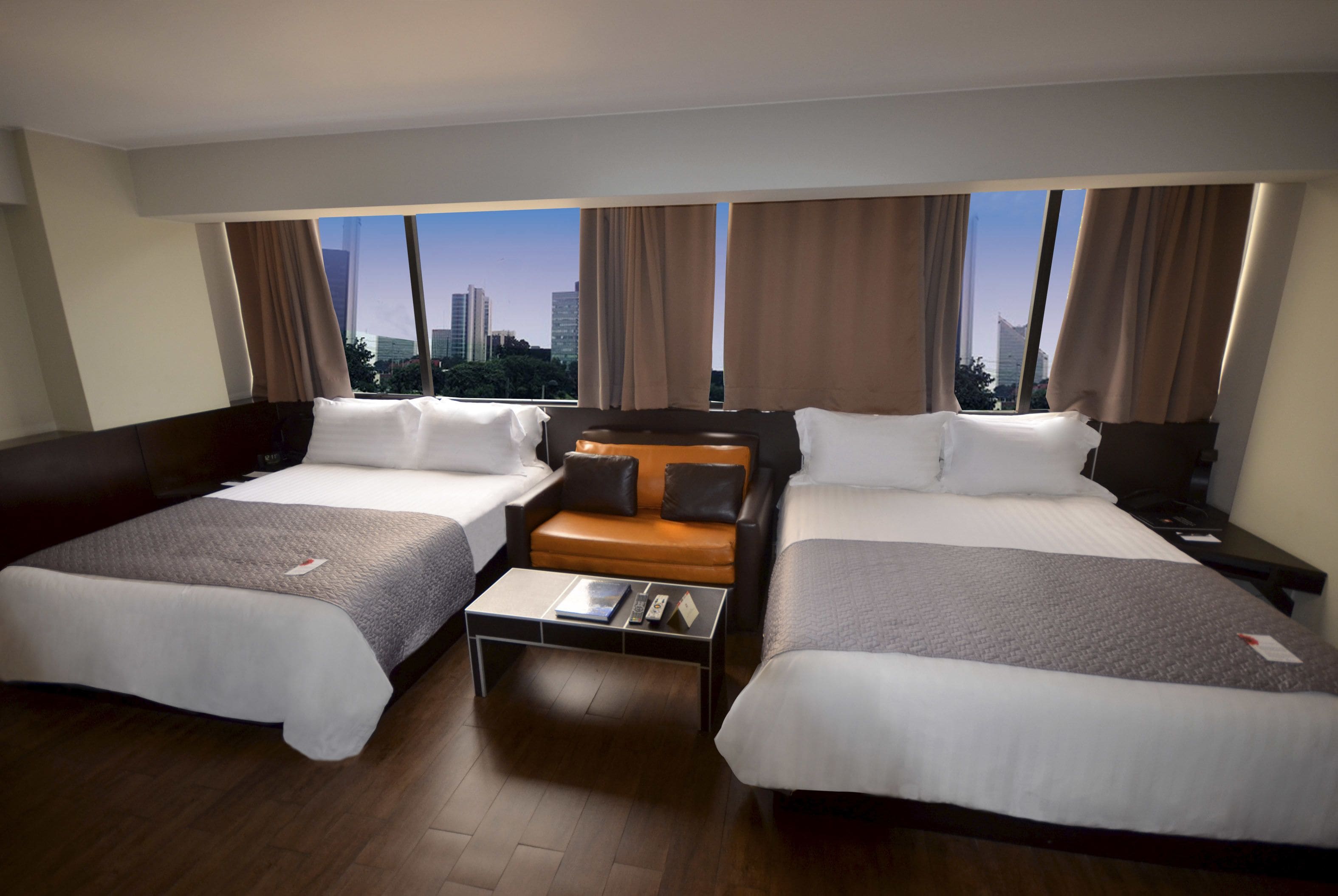 Quarto de hotel com duas camas, um sofá pequeno, uma mesa de centro e vista para a paisagem urbana.