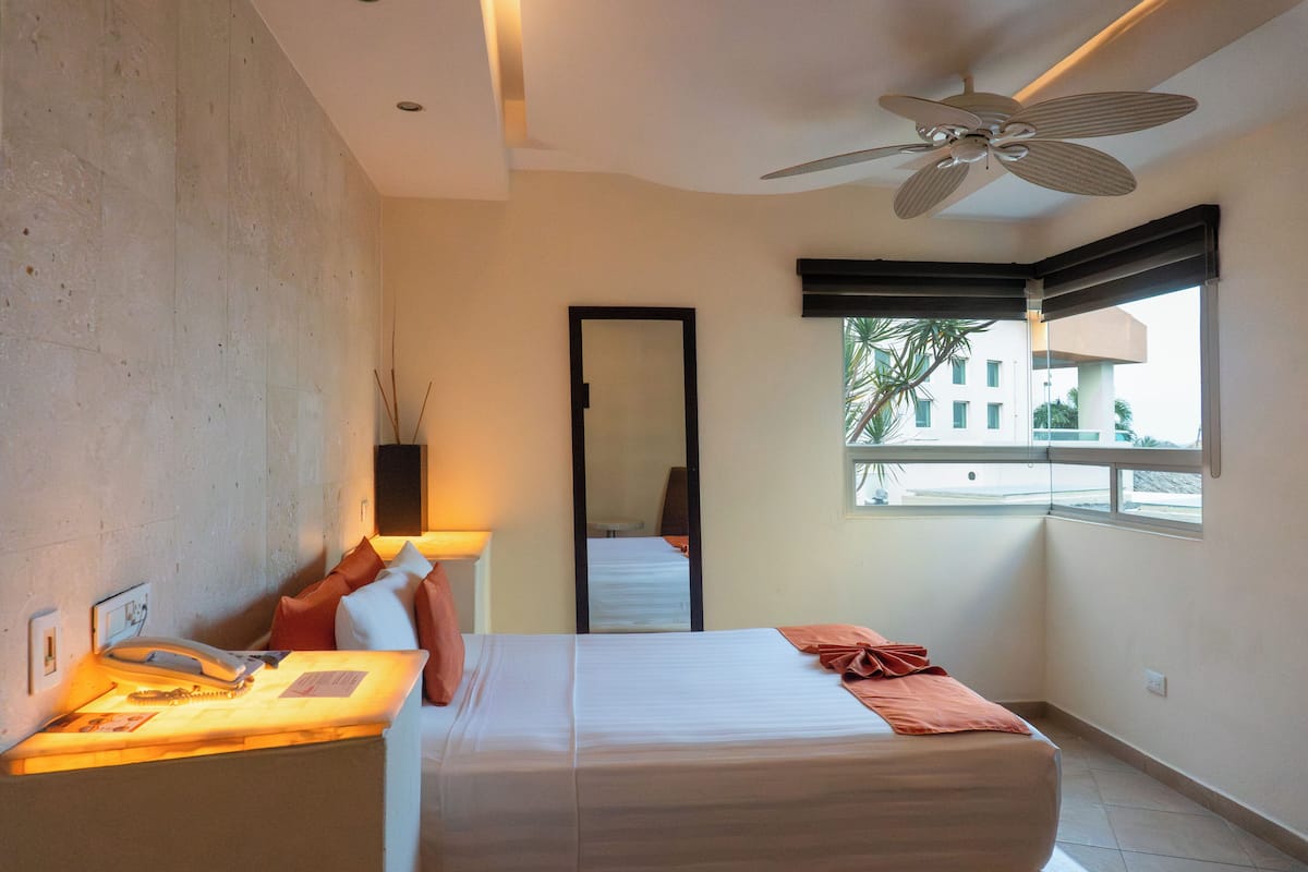 Habitación de hotel con cama, escritorio con lámpara, ventilador de techo, ventana grande y vista al exterior.