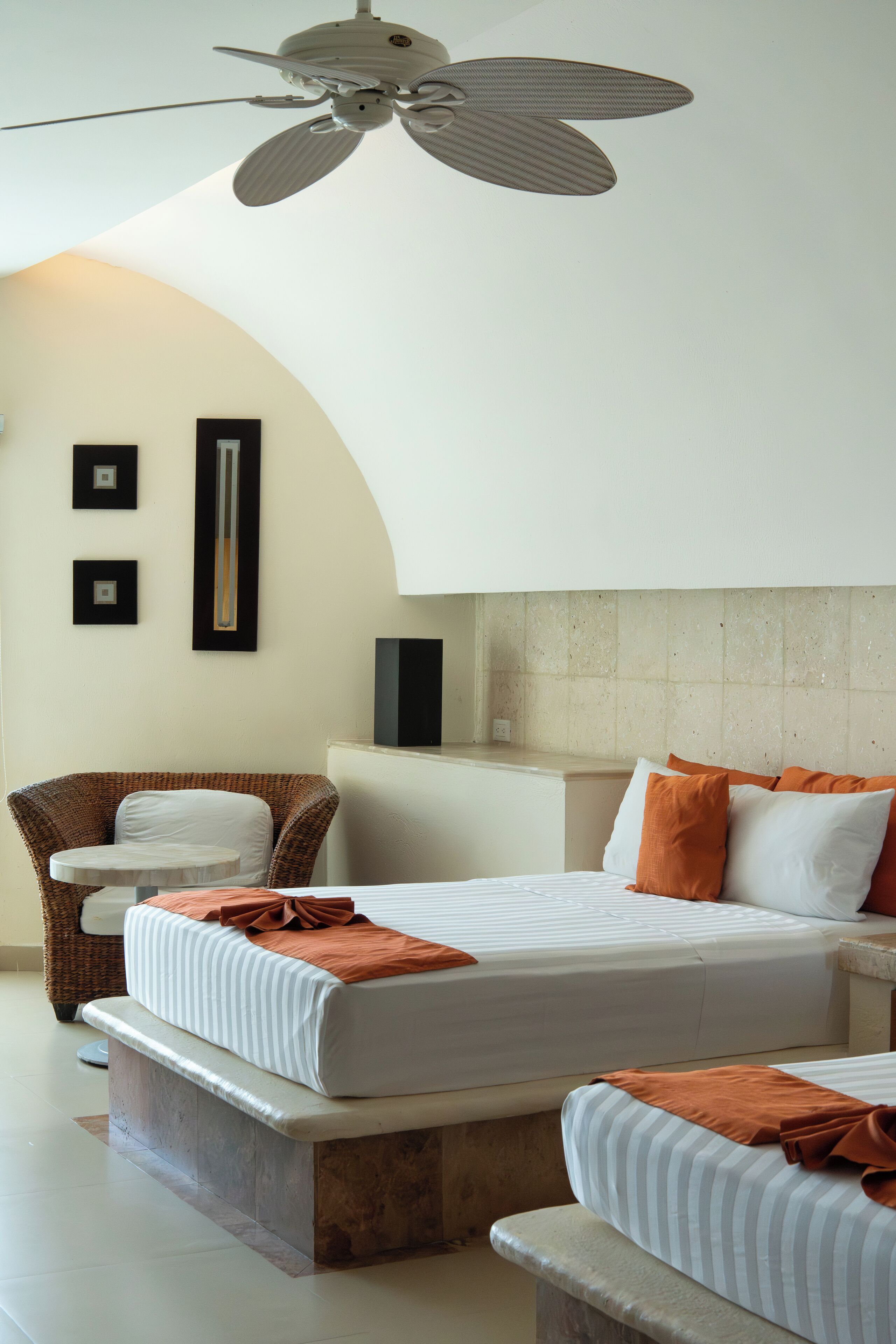 Habitación de hotel con dos camas, ventilador de techo, una silla de mimbre y una mesita.