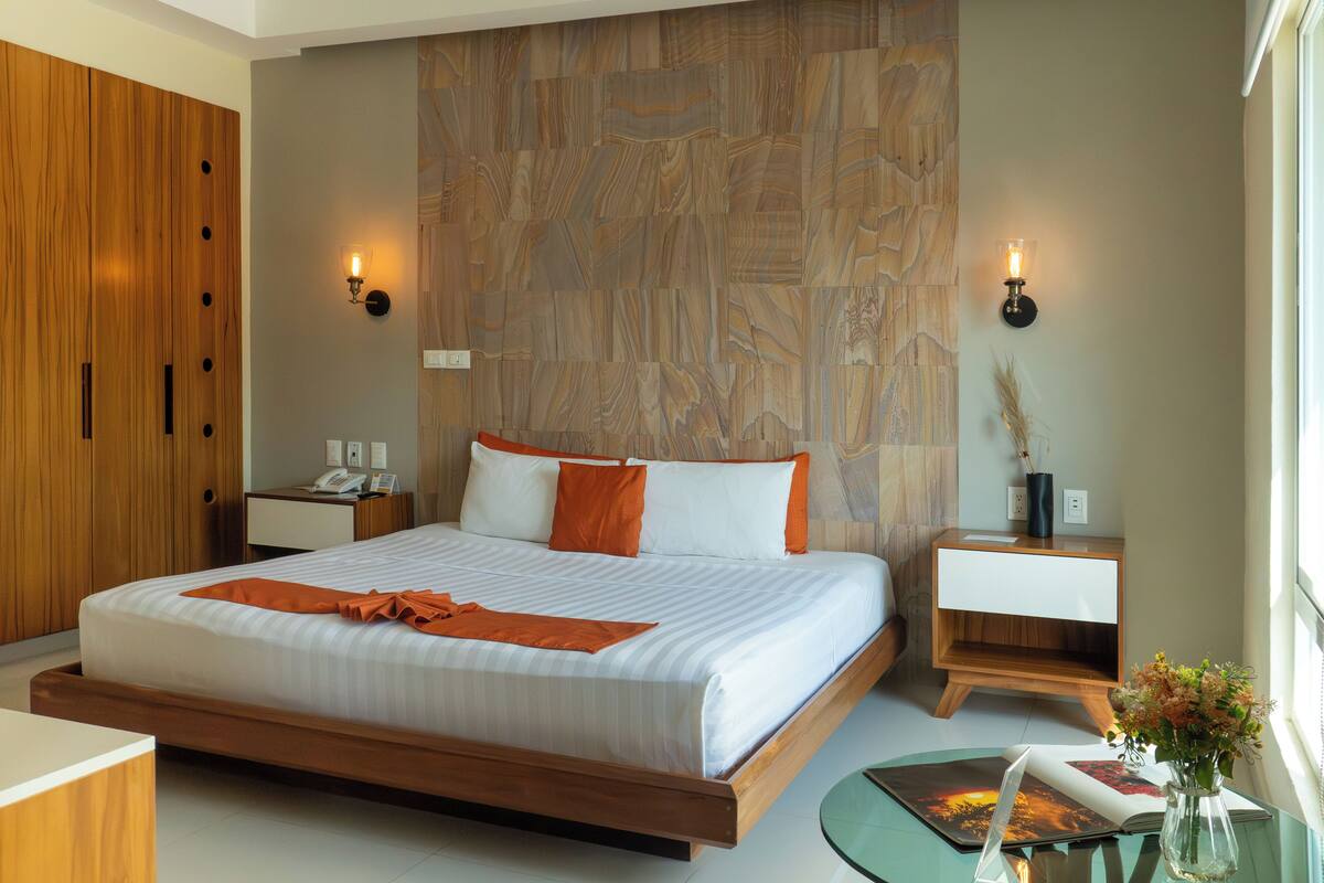 Una habitación de hotel moderna con una cama grande, un armario de madera, mesitas de noche y una mesita auxiliar.
