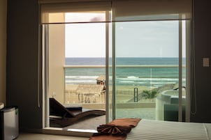 Habitación con un ventanal grande con vista a una playa y al océano, una cama con almohada y una mesita.