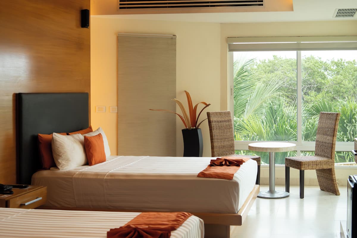 Habitación de hotel con dos camas, una mesita, sillas de mimbre, una planta en maceta y un ventanal con vistas a la vegetación.