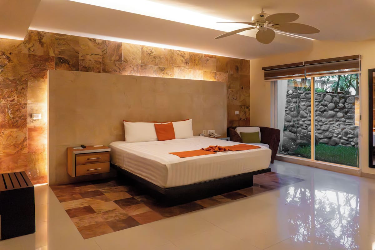 Una habitación de hotel moderna con una cama grande, una mesita de noche, un escritorio y vistas a un muro de piedra y follaje exterior.