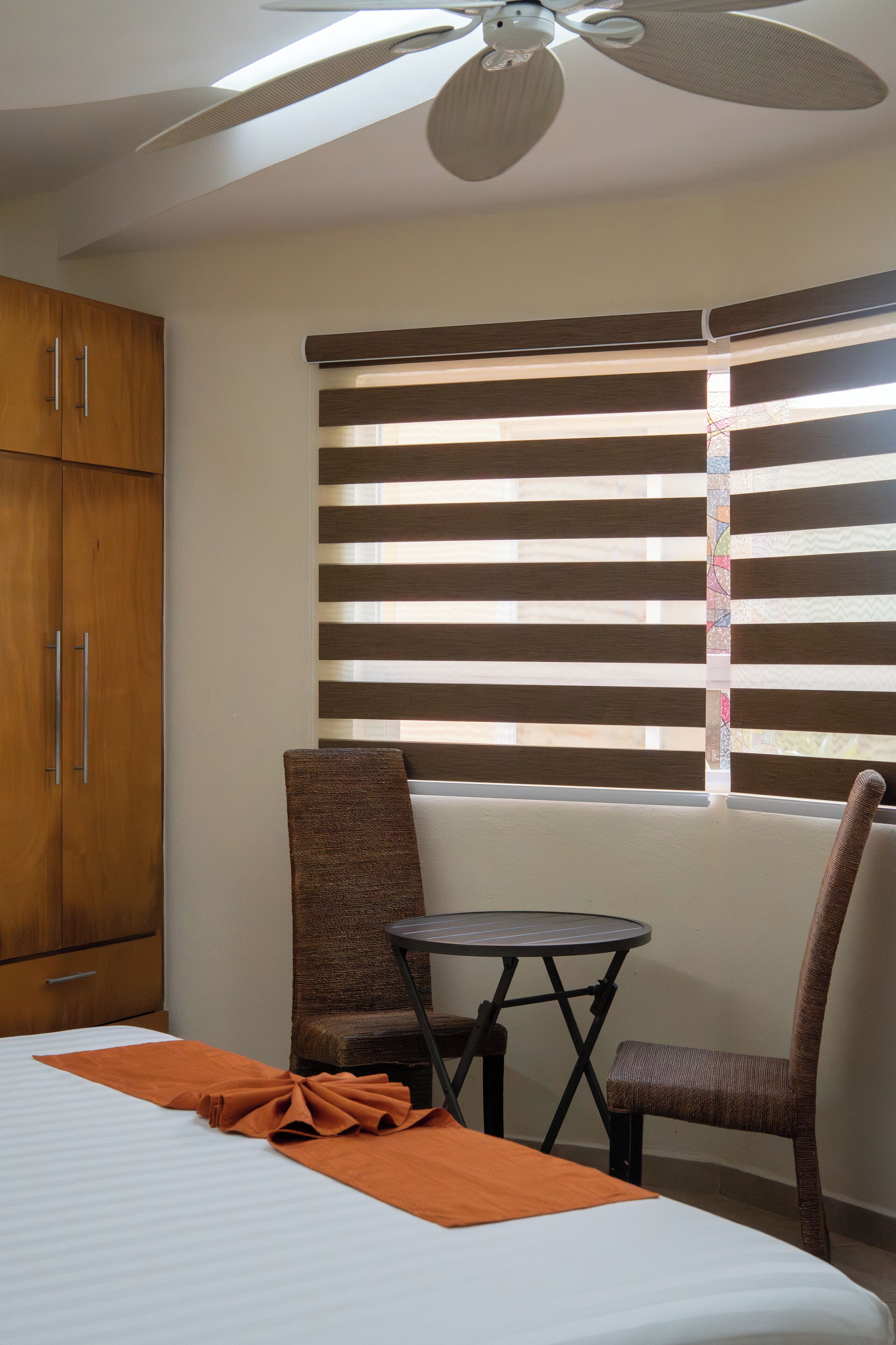 Habitación de hotel con cama, una mesita, dos sillas, ventilador de techo y ventana con persianas.