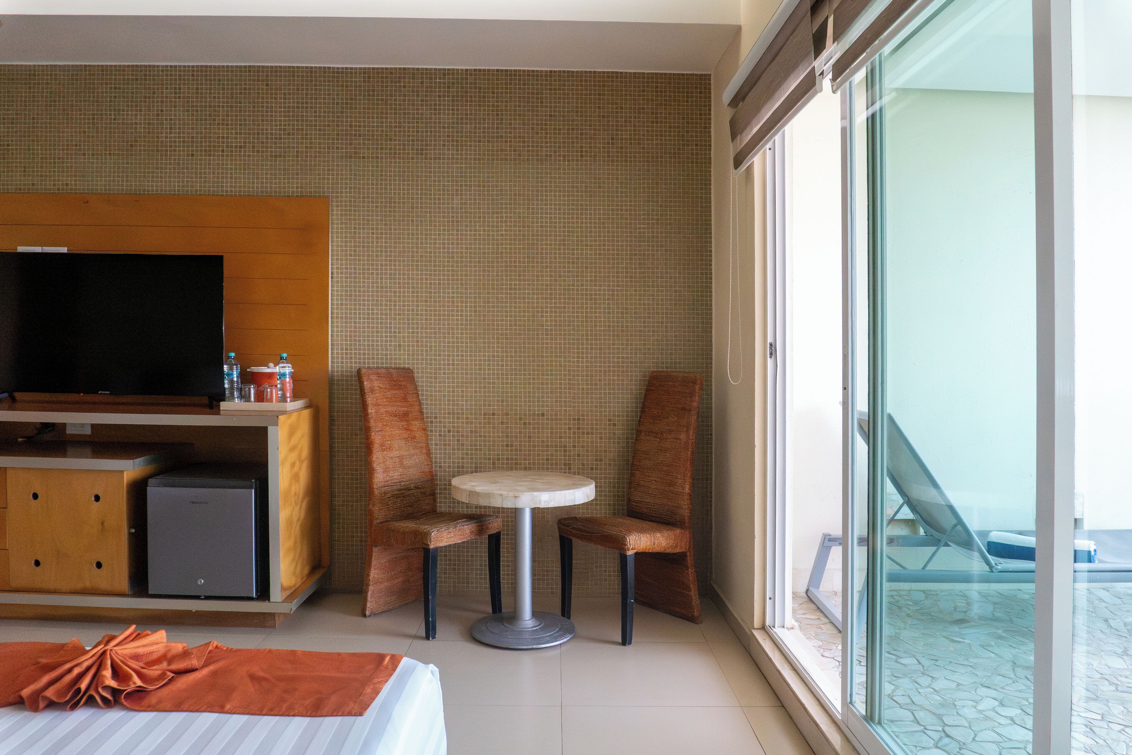 Une chambre d’hôtel comprenant un lit, une télévision, un petit réfrigérateur, une table avec deux chaises et un balcon avec un fauteuil.