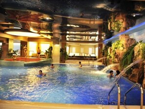 An indoor swimming pool with a waterfall feature, lounge areas, and a view of the sky.