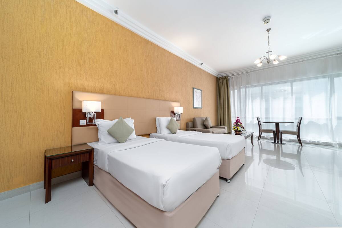A hotel room with two beds, a small table, a chair, and a large window with curtains.
