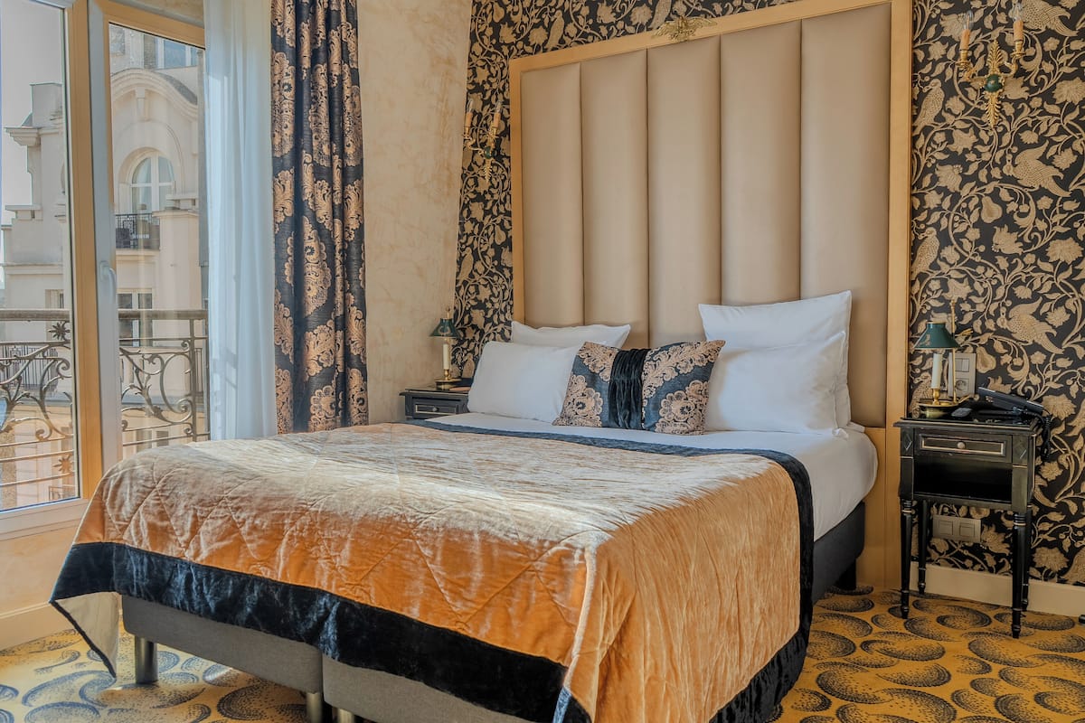 A hotel room with a large bed, a nightstand, a lamp, and a view of the city through a window.