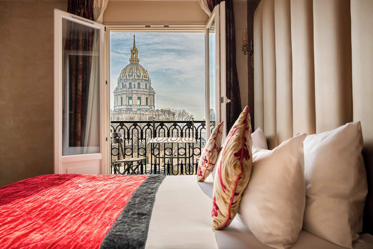 A hotel room with a bed, pillows, and a view of a large building through an open window.