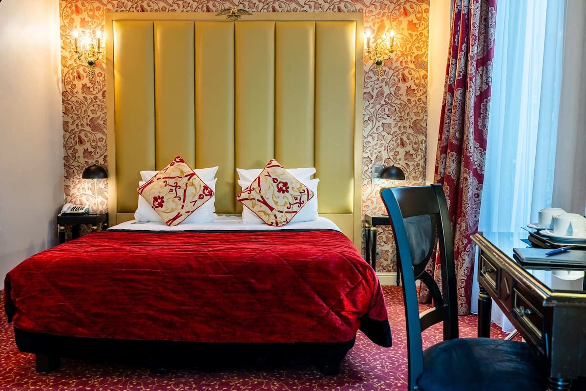 A hotel room with a large bed, two decorative pillows, a nightstand with a lamp, a desk with a chair, and a window with curtains.