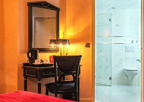A hotel room with a bed, a desk with a lamp, a chair, a mirror, and a bathroom with a toilet and shower.