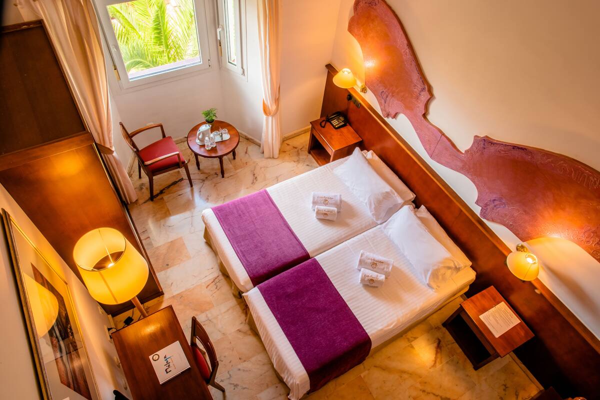 A hotel room with two beds, a desk, a chair, a small table with a plant, and a window with a view of greenery.