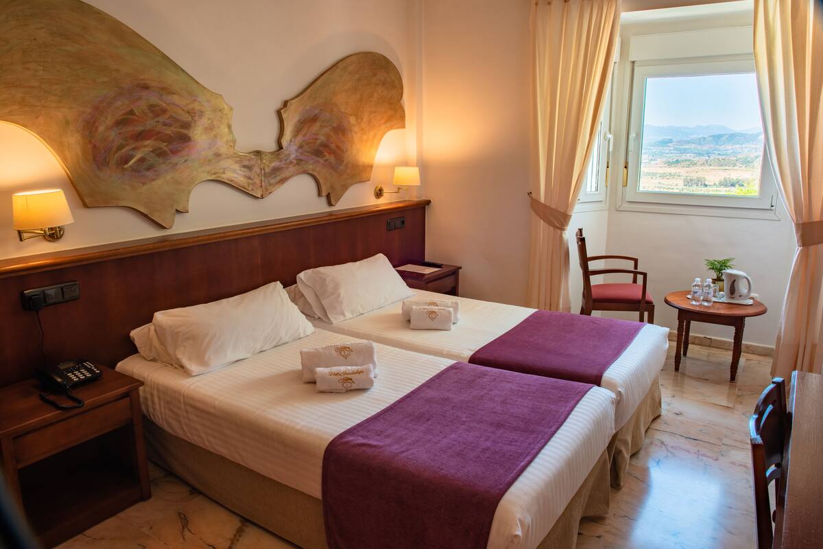 A hotel room with two beds, a wooden headboard, a nightstand with a phone, a small table with a plant, and a window with curtains.