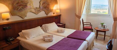 A hotel room with two beds, a wooden headboard, a nightstand with a phone, a small table with a plant, and a window with curtains.