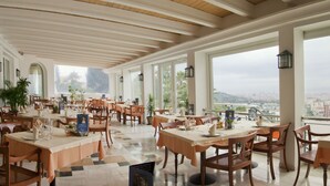 Een ruime restaurantruimte met meerdere tafels en stoelen, grote ramen met uitzicht op de stad en een hoog plafond met houten balken.