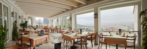 Un restaurante espacioso con numerosas mesas y sillas, grandes ventanales que ofrecen vistas a la ciudad y un techo alto con vigas de madera.