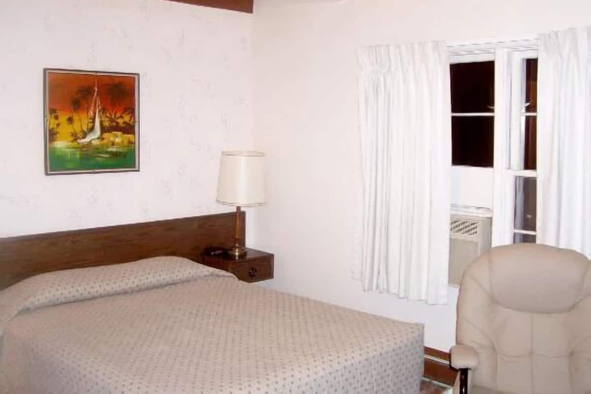 A hotel room with a bed, a nightstand with a lamp, a window with curtains, and a painting on the wall.