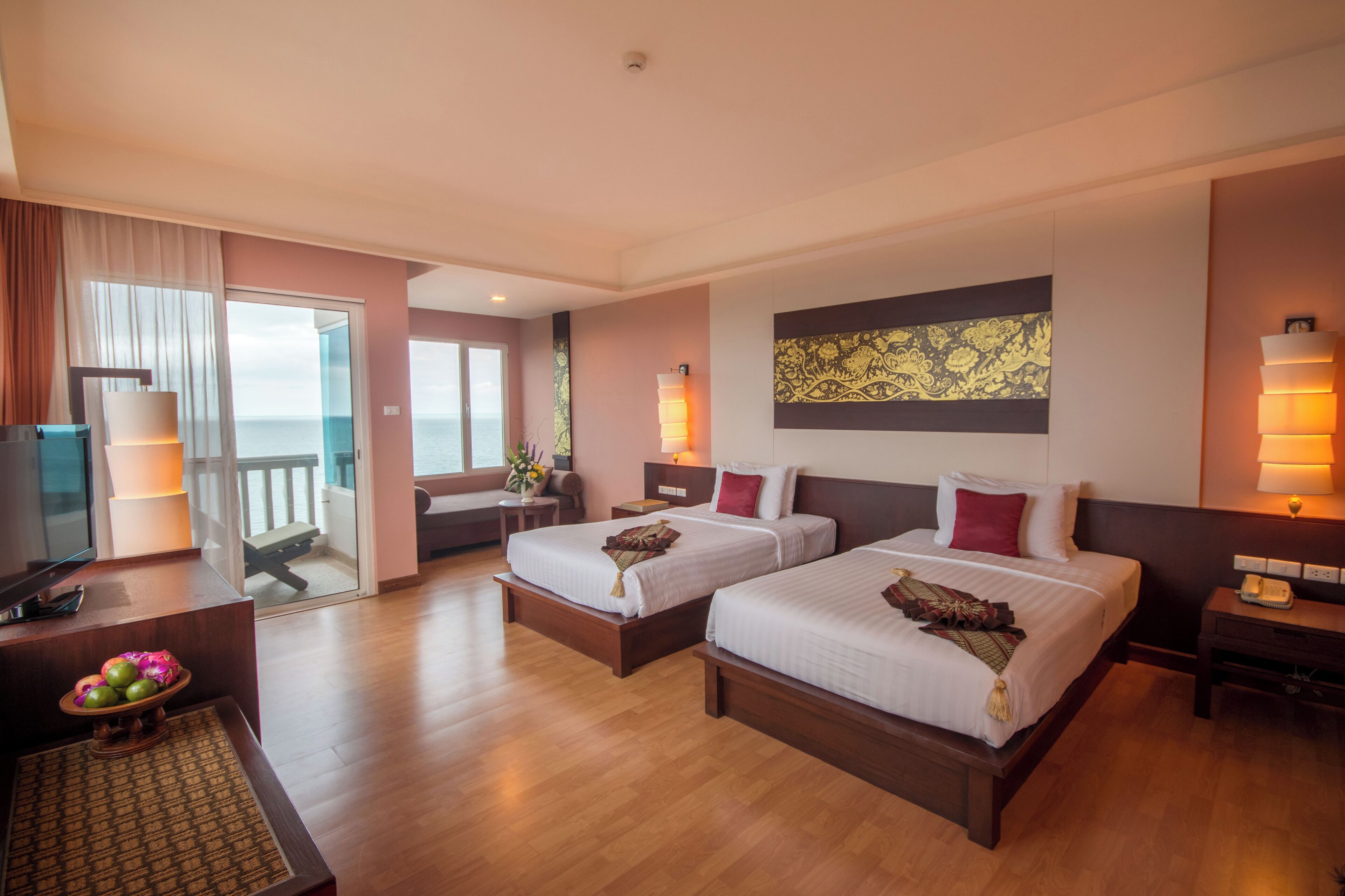 Quarto de hotel com duas camas, televisão, escrivaninha com luminária e vista para o mar.