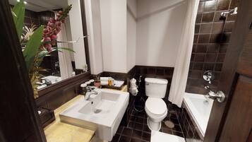 Un baño con lavamanos, inodoro y bañera blancos. Hay un espejo sobre el lavamanos y una planta a la izquierda.