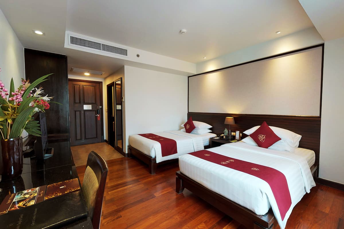 Habitación de hotel con dos camas, un escritorio, una silla, un florero con flores y piso de madera.