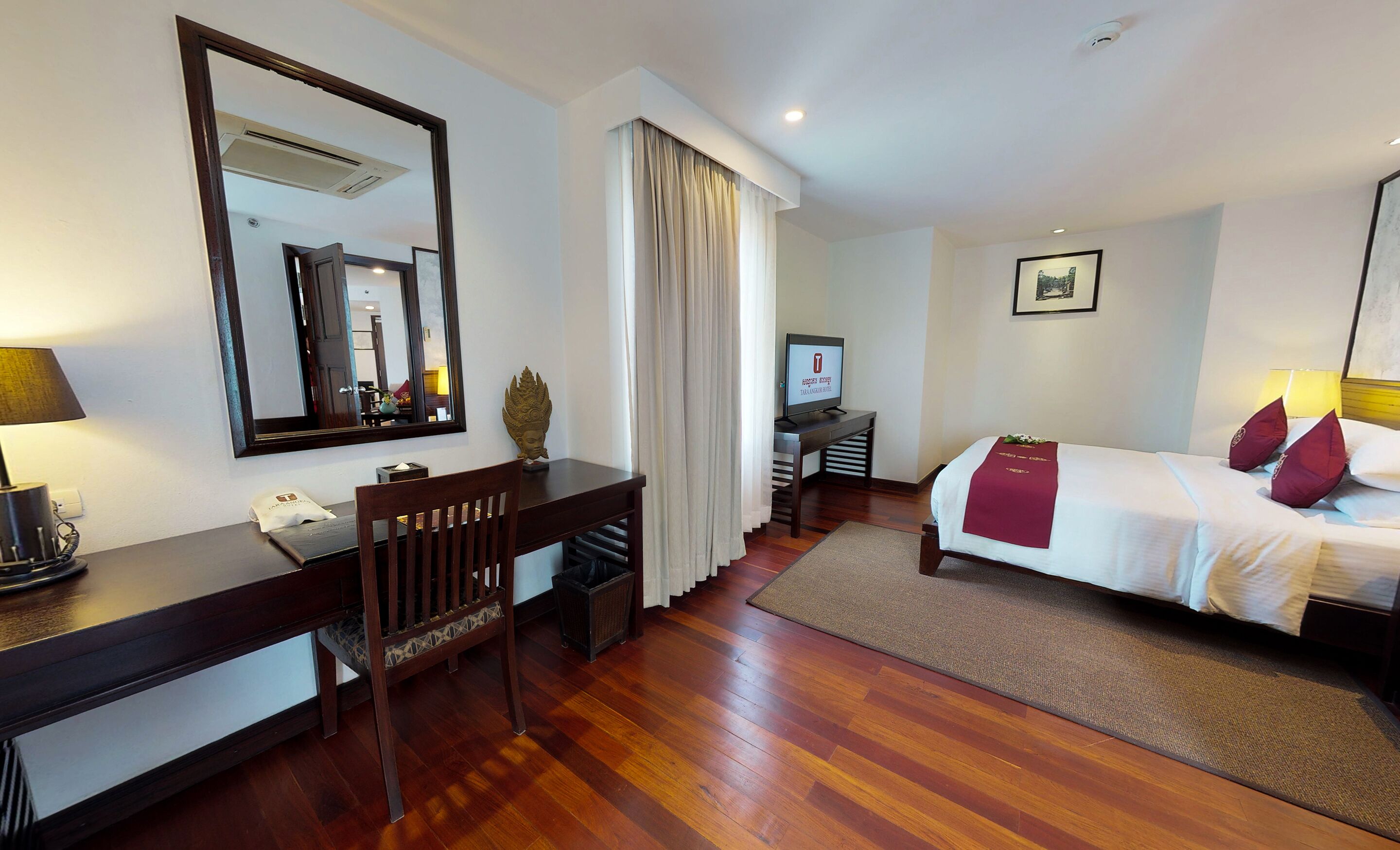 A hotel room with a wooden desk, a bed with red pillows, a TV, and a large mirror.