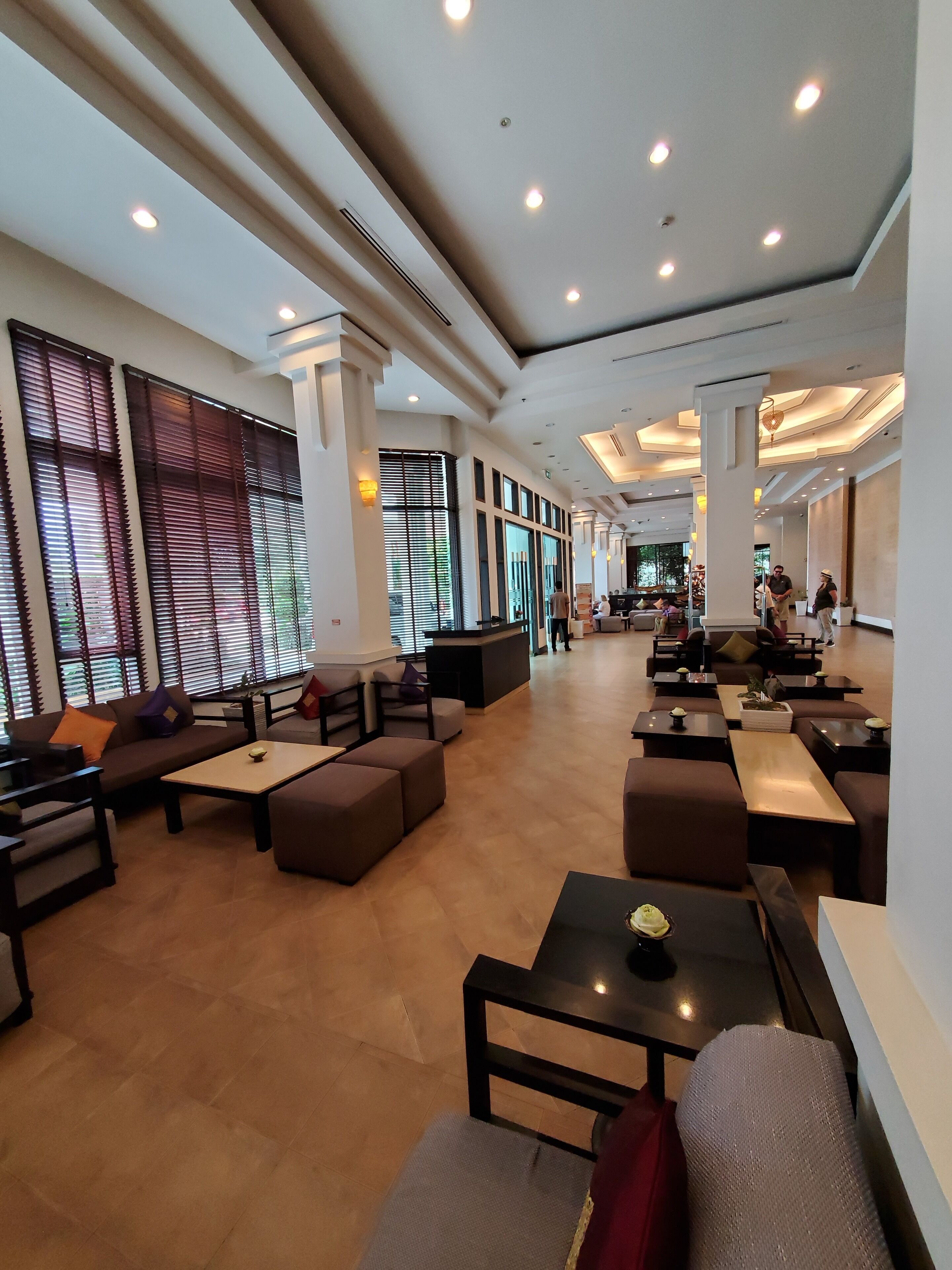 A spacious lobby with multiple seating areas, large windows with blinds, and a modern ceiling design.