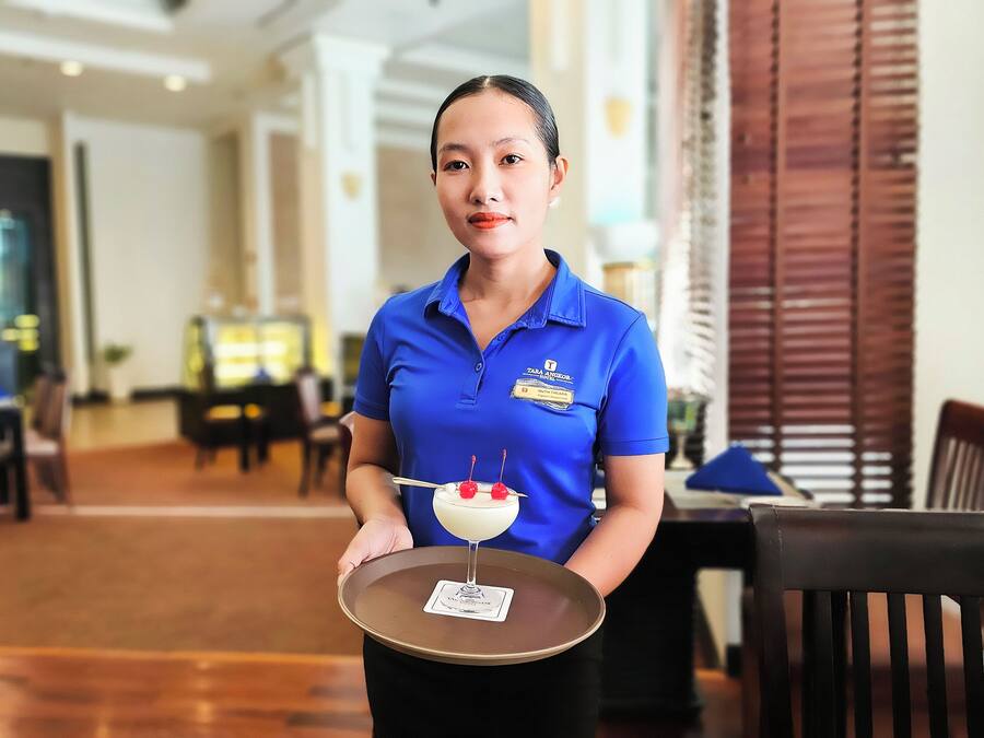 A hotel staff member holding a tray with a dessert. The setting appears to be a dining area with tables and chairs.
