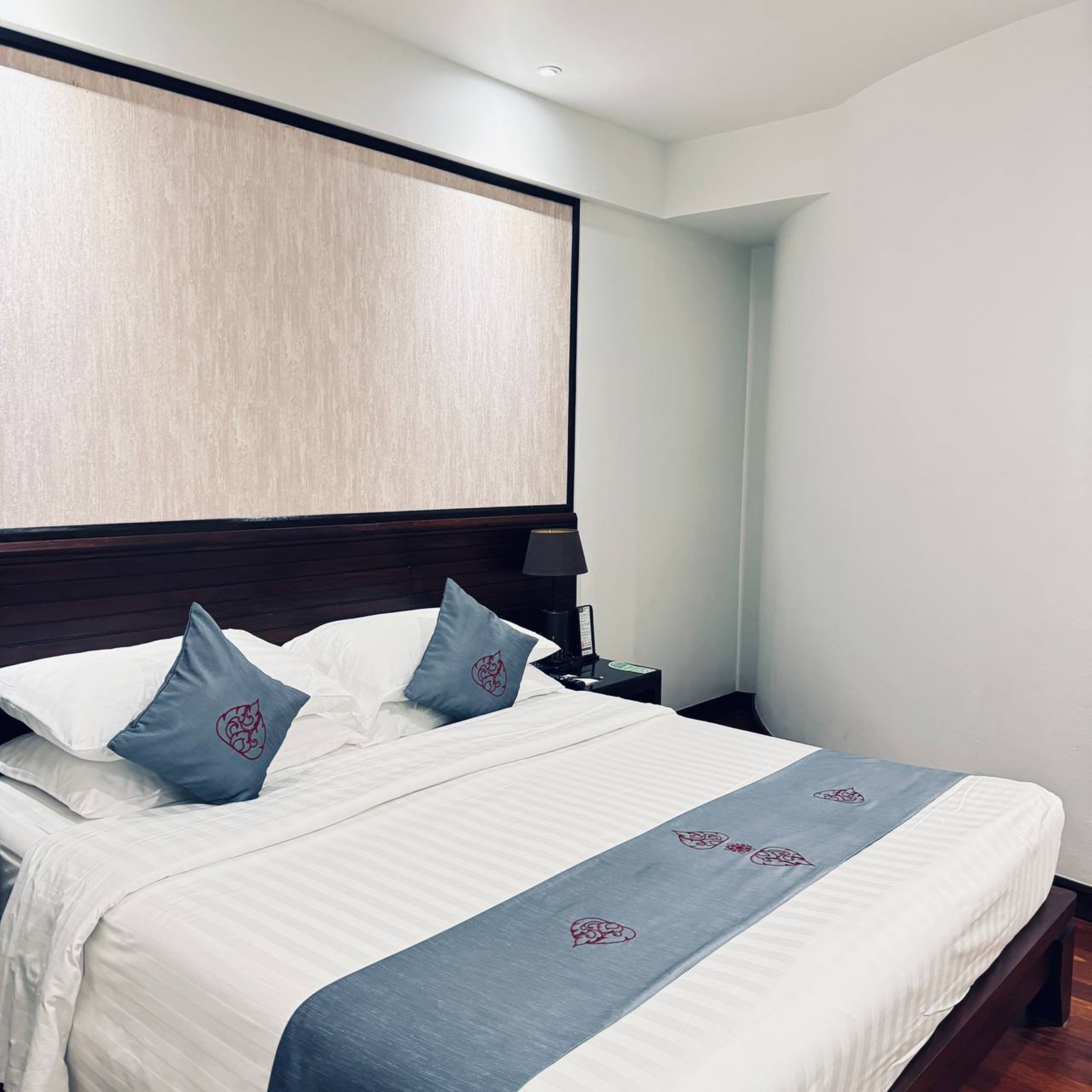 A neatly made bed with white linens and blue accents, a dark wooden headboard, and a nightstand with a lamp.
