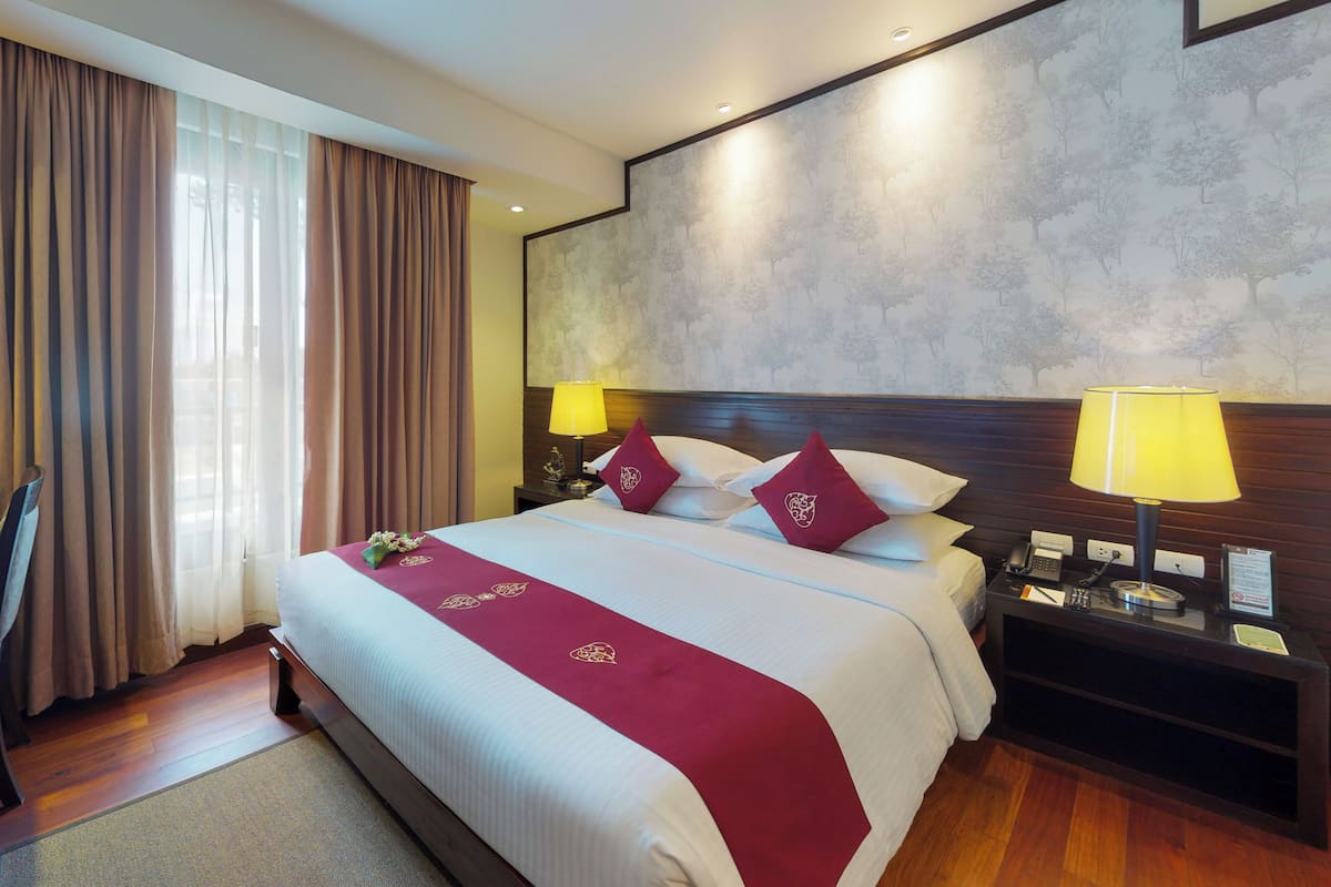 Habitación de hotel con una cama grande, dos lámparas de noche, un cabecero de madera, un escritorio con una silla y vistas a árboles a través de la ventana.
