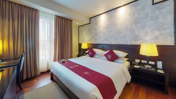 Une chambre d’hôtel avec un grand lit, deux lampes de chevet, une tête de lit en bois, un bureau avec une chaise et une vue sur les arbres par la fenêtre.
