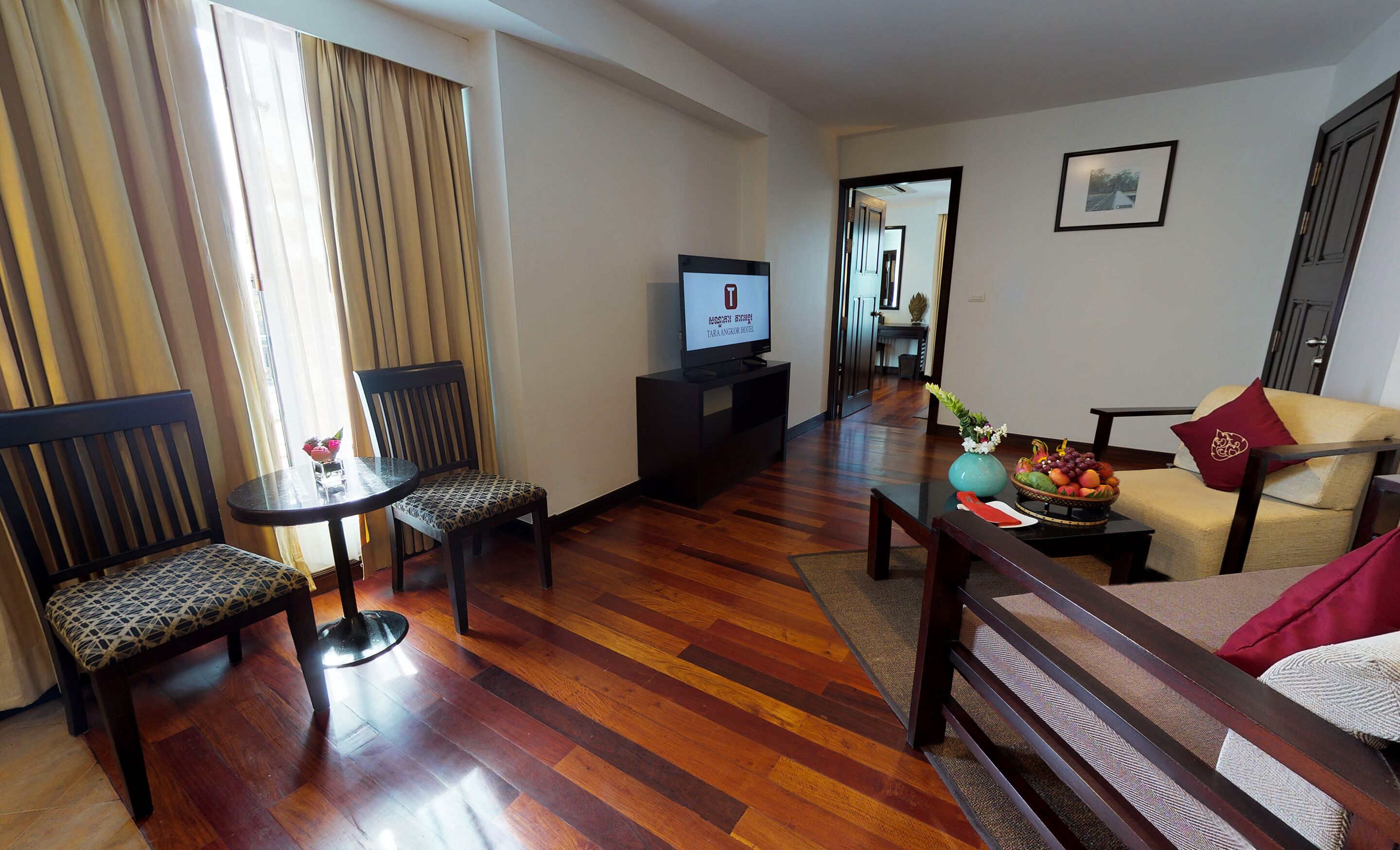 Une chambre d’hôtel avec un plancher en bois, un téléviseur posé sur un meuble, deux chaises, un canapé et un panier de fruits.