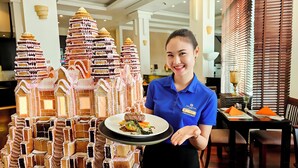 A hotel staff member holding a tray with a dessert, standing in front of a gingerbread house display. - Tara Angkor Hotel (Siem Reap)