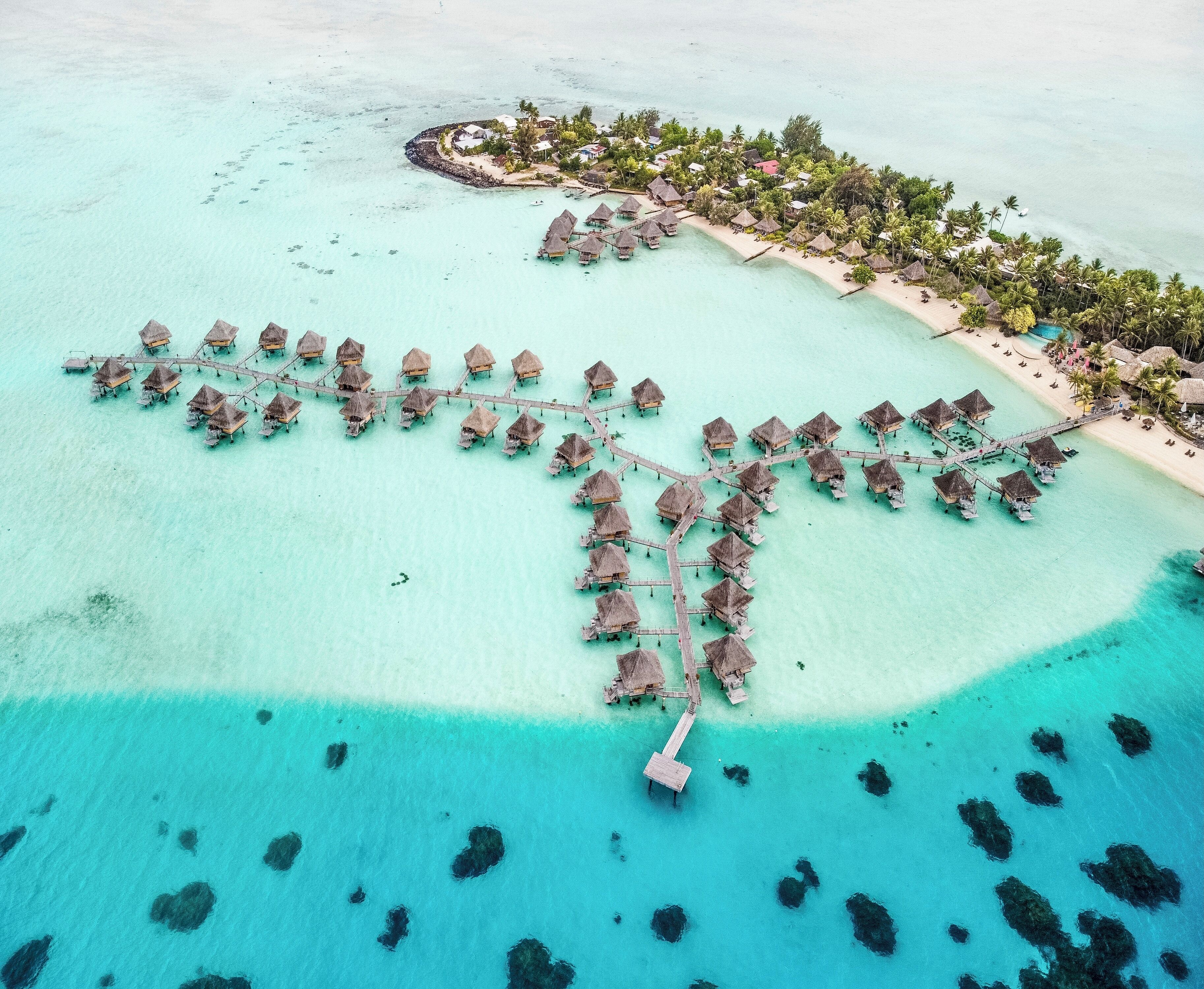 Vista aérea de un resort con bungalows sobre el agua unidos por pasarelas, aguas turquesas cristalinas y una pequeña isla con estructuras adicionales.
