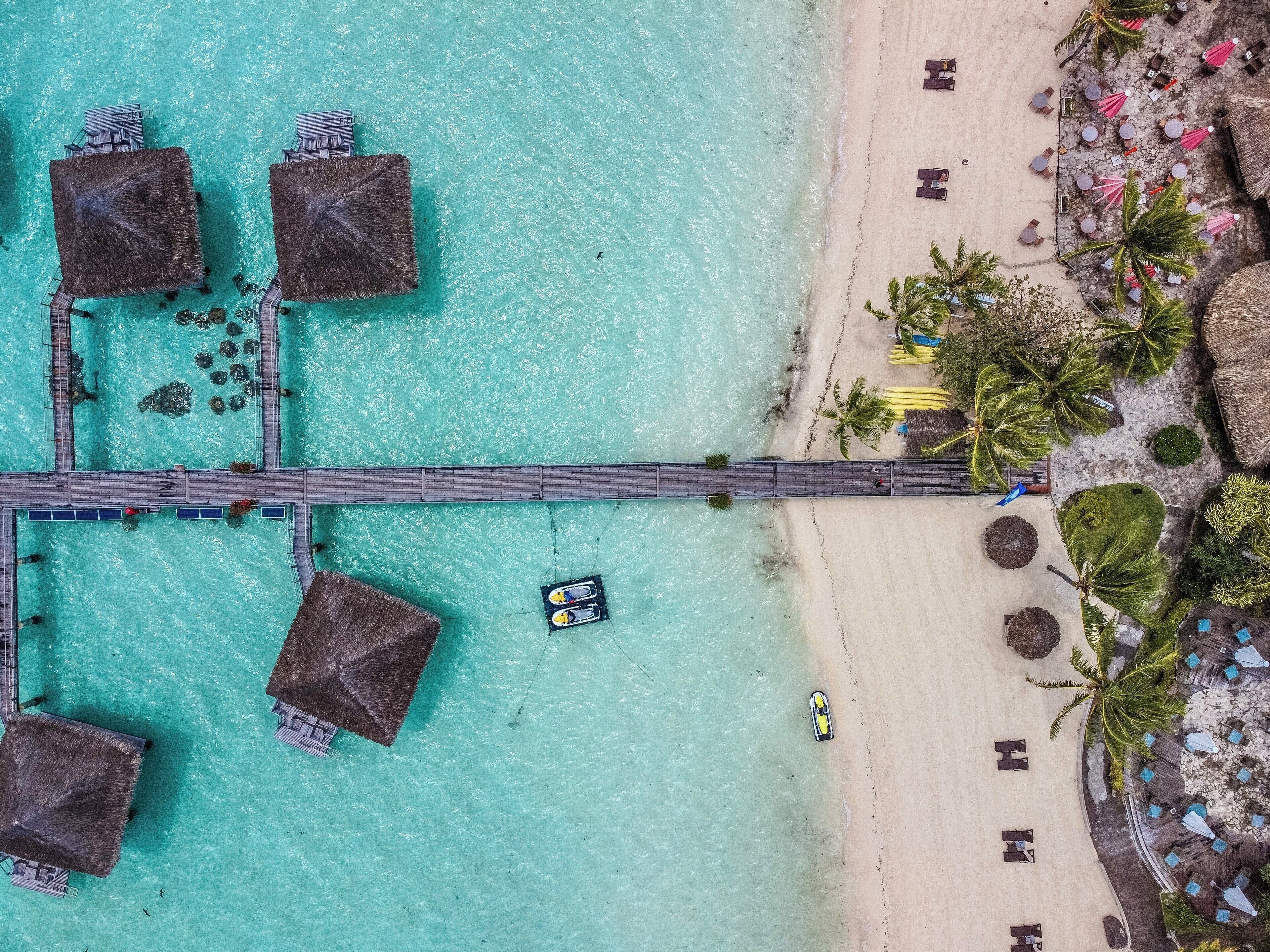 An aerial view of a beach with overwater bungalows, a pier, and beach umbrellas.