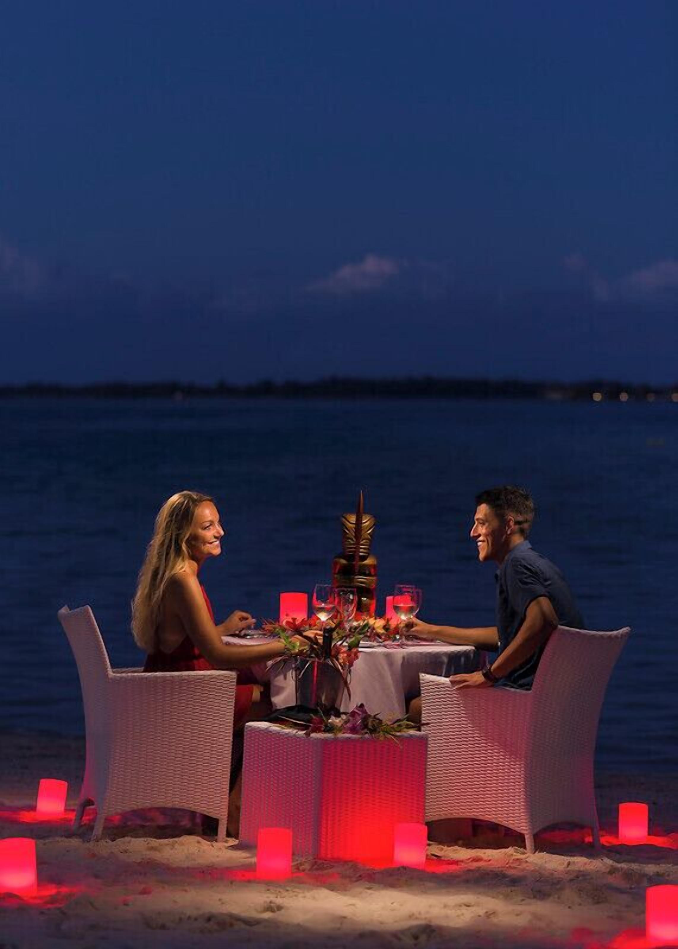 A dining setup with a table and chairs on a beach at night, overlooking the sea.