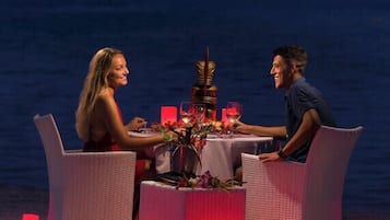 A dining setup with a table and chairs on a beach at night, overlooking the sea.