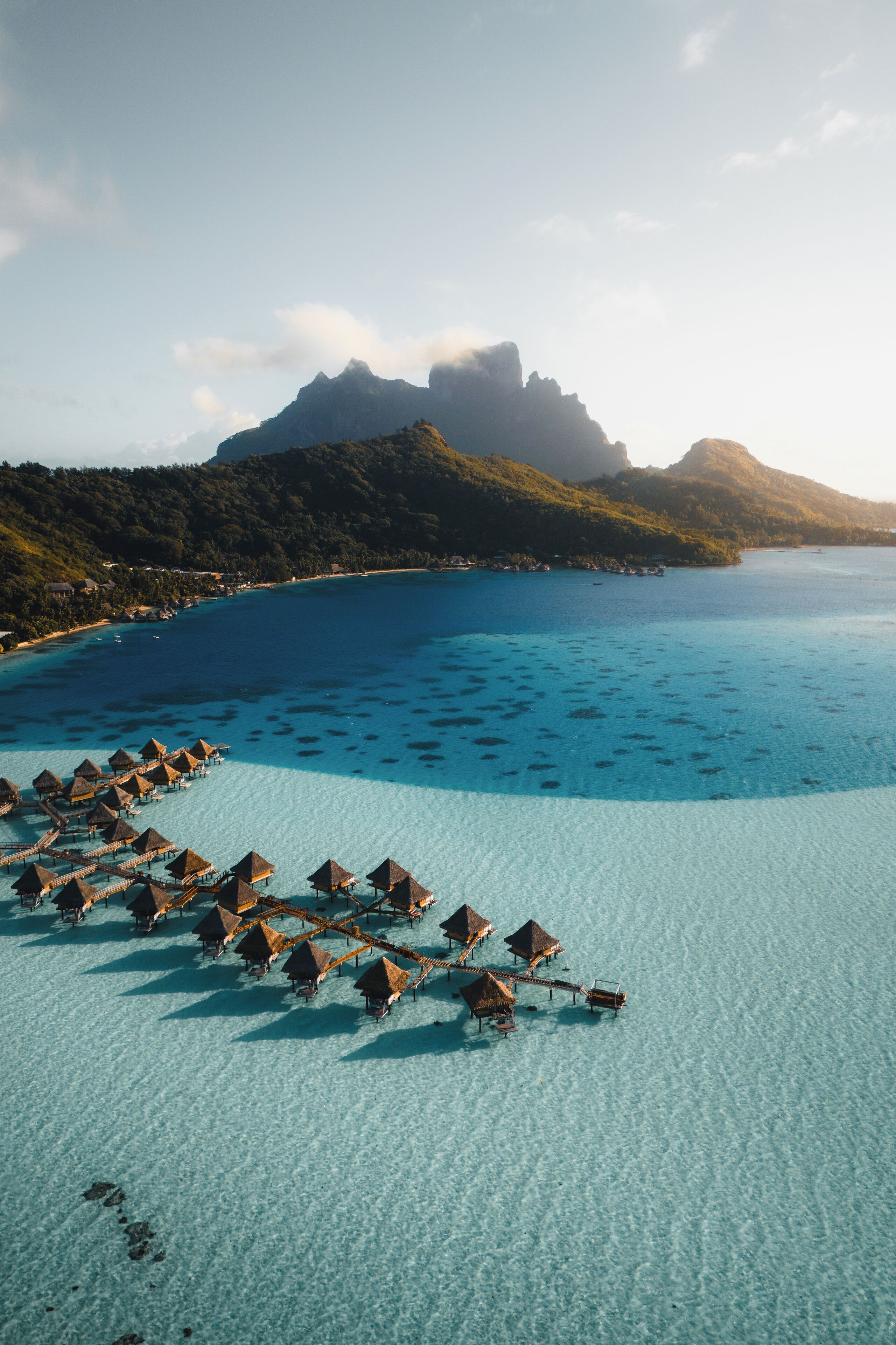 An aerial view of overwater bungalows on a clear turquoise sea, surrounded by lush greenery and a prominent mountain in the background.