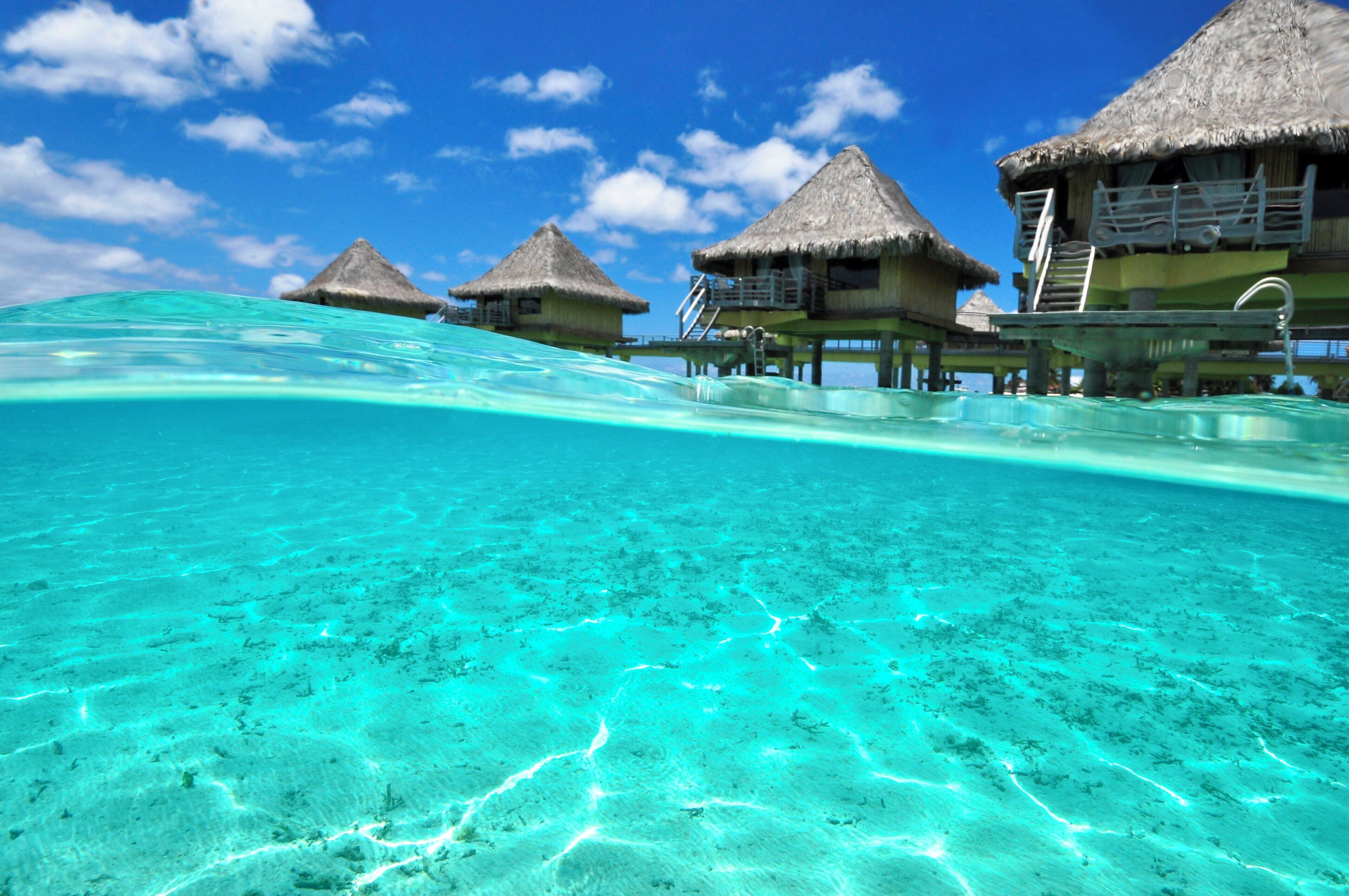 An underwater view of a clear turquoise sea with overwater bungalows featuring thatched roofs and wooden walkways.