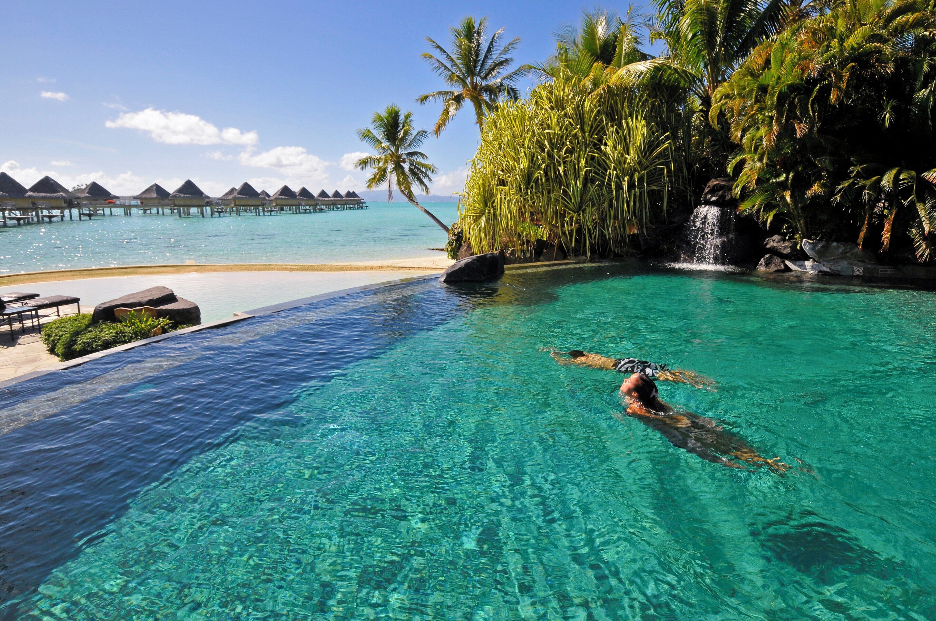 Piscina azul cristalina con una persona nadando, rodeada de vegetación tropical y vistas a bungalows sobre el agua.