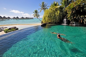 A clear blue pool with a person swimming, surrounded by tropical vegetation and a view of overwater bungalows.