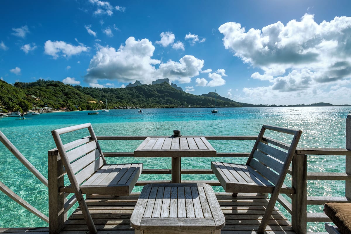 Eine Holzterrasse mit zwei Stühlen und einem Tisch mit Blick auf türkisfarbenes Wasser, wo Boote fahren und eine Berglandschaft im Hintergrund liegt.