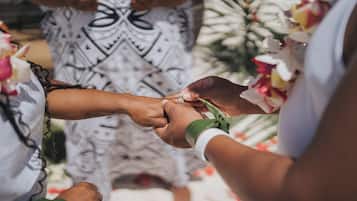 A close-up of hands exchanging rings, with a patterned garment and floral decorations in the background.