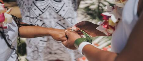 A close-up of hands exchanging rings, with a patterned garment and floral decorations in the background.