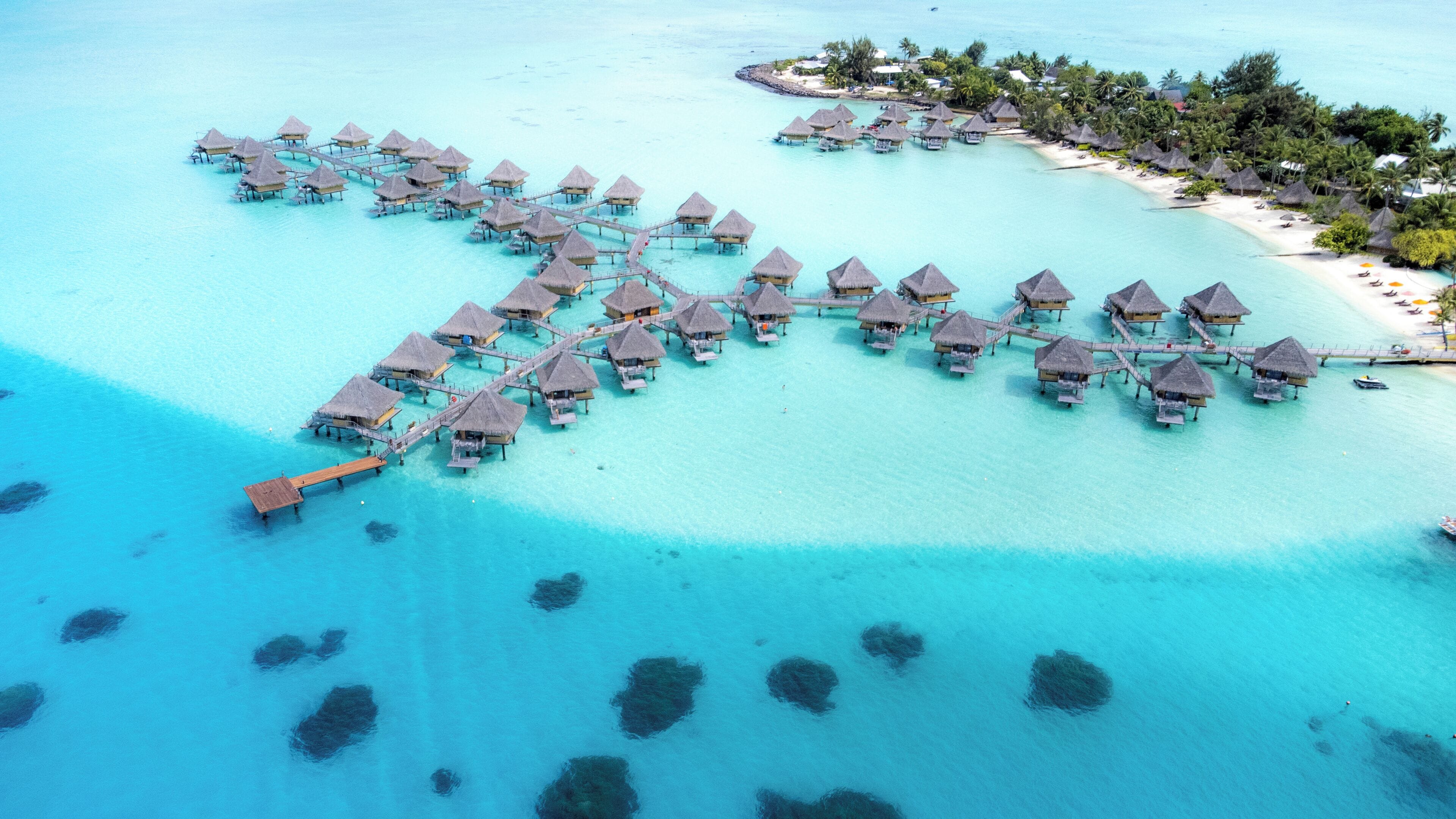 Vista aérea de bungalows sobre el agua unidos por senderos, rodeados de aguas turquesas cristalinas y una pequeña isla con palmeras.