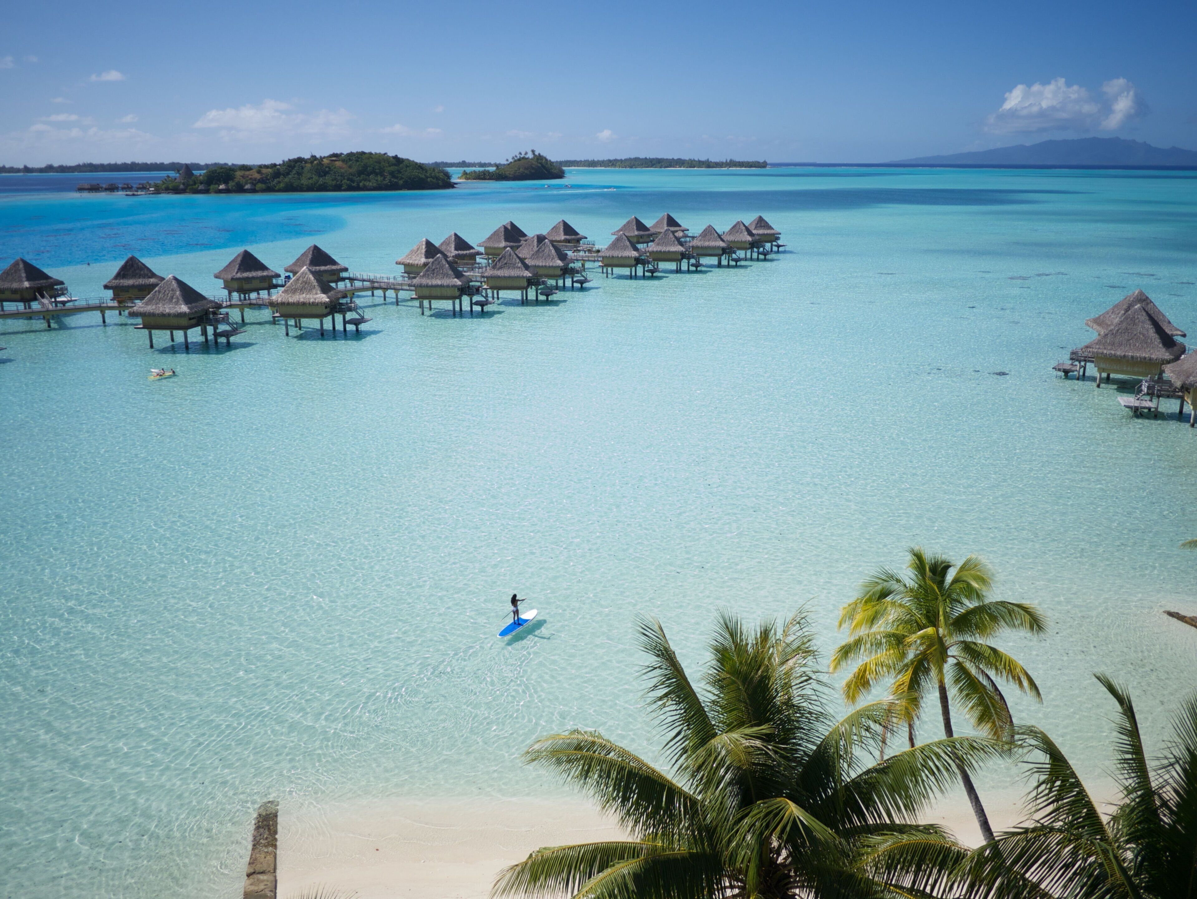 Overwater bungalows with clear turquoise water, a sandy beach, and palm trees.