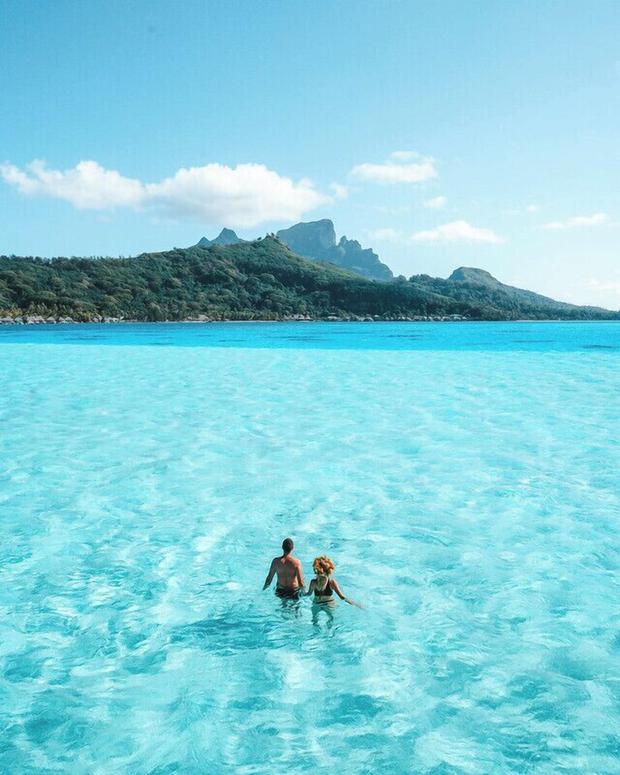 A couple walking in clear turquoise water with a mountainous landscape in the background.