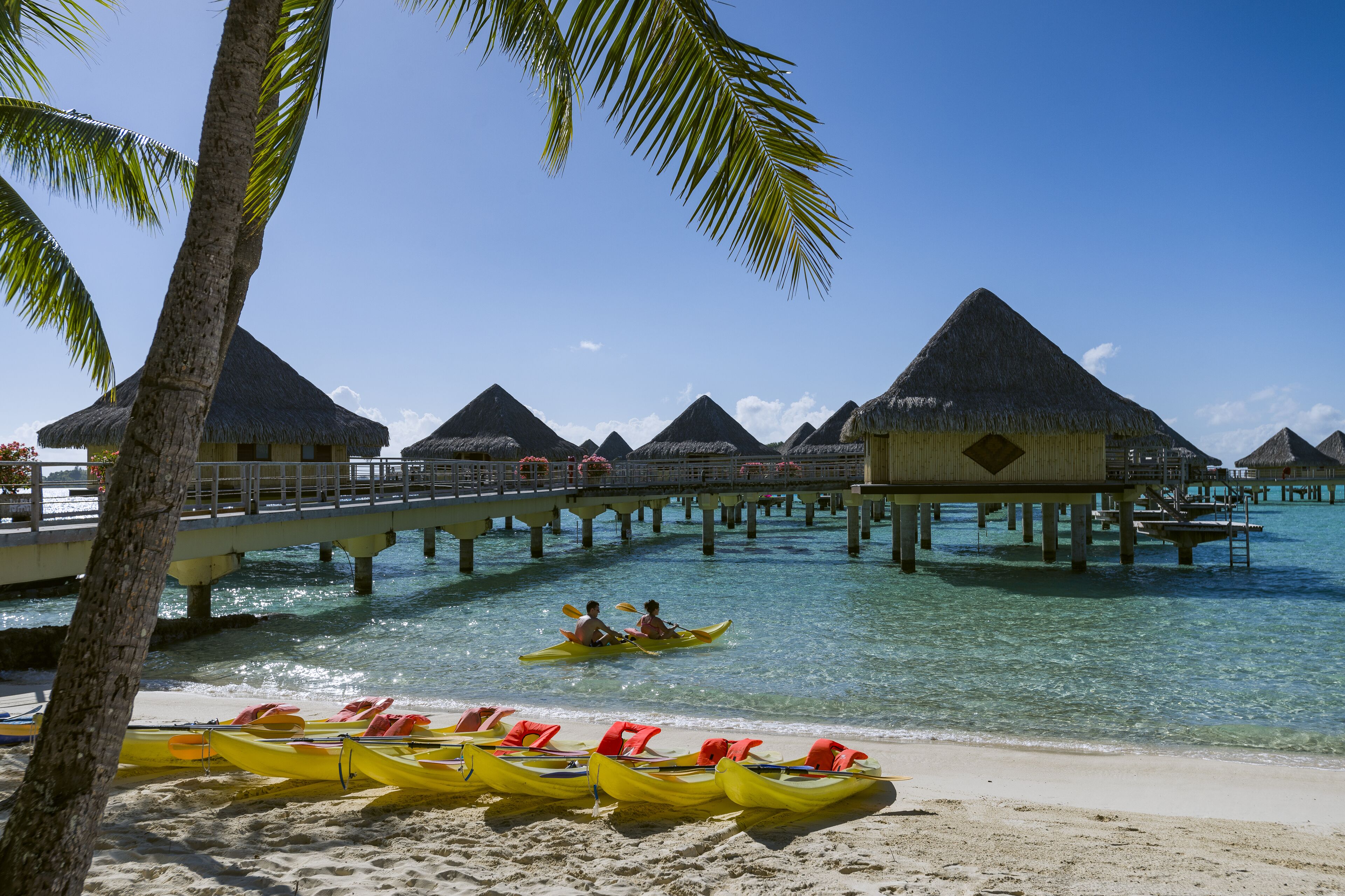 Une plage avec des bungalows sur pilotis, des kayaks et un pont.