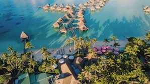 Eine Luftaufnahme eines Resorts mit überwasserbungalows, die durch Wege miteinander verbunden sind, einem Strandabschnitt mit Sonnenschirmen und Palmen.