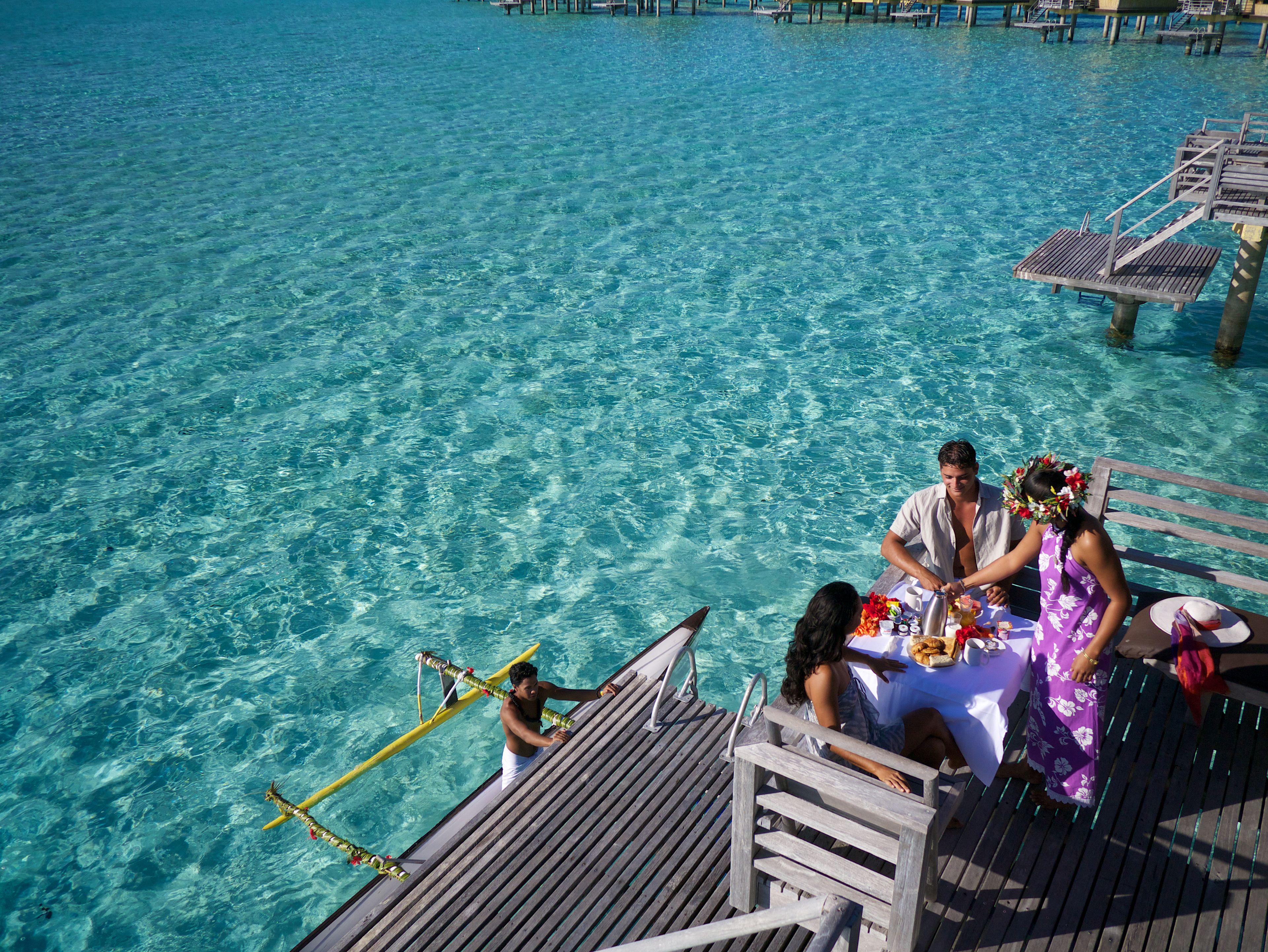 A wooden deck with a table set for two, overlooking clear turquoise water.