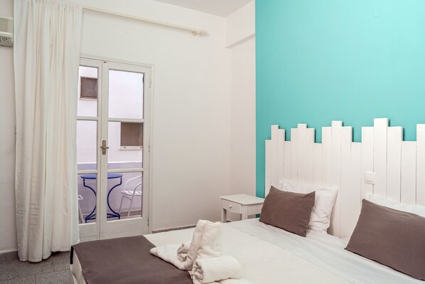 A hotel room with a bed, a small bedside table, a window with curtains, and a view of the outside balcony. - Ikaros Hotel (Santorini)